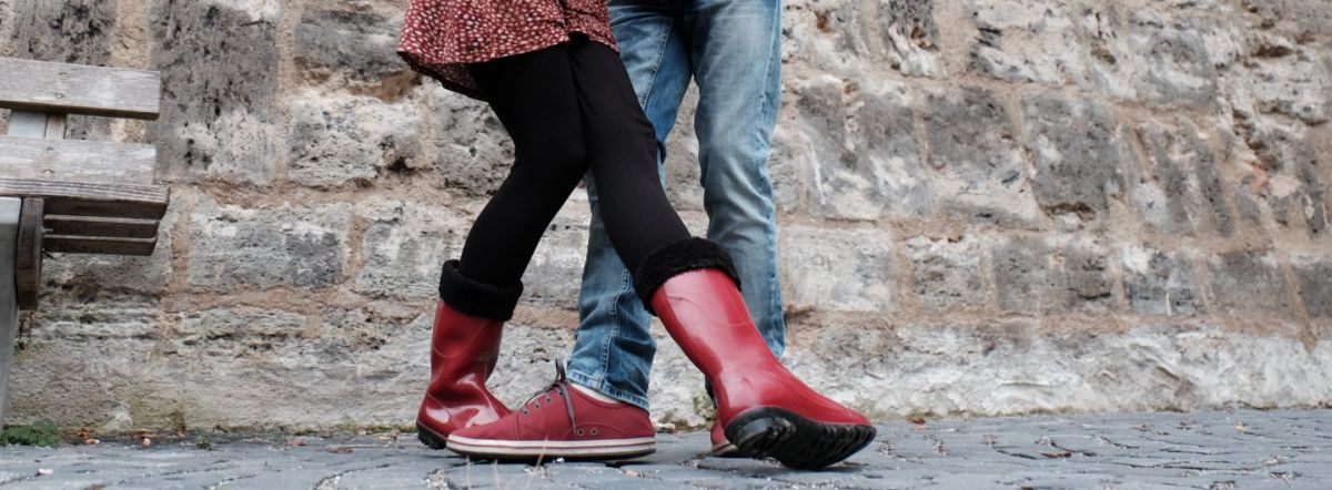 rote Gummistiefel und rote Sneaker in verschränkter Tanzposition vor Stadtmauer und neben einer Sitzbank