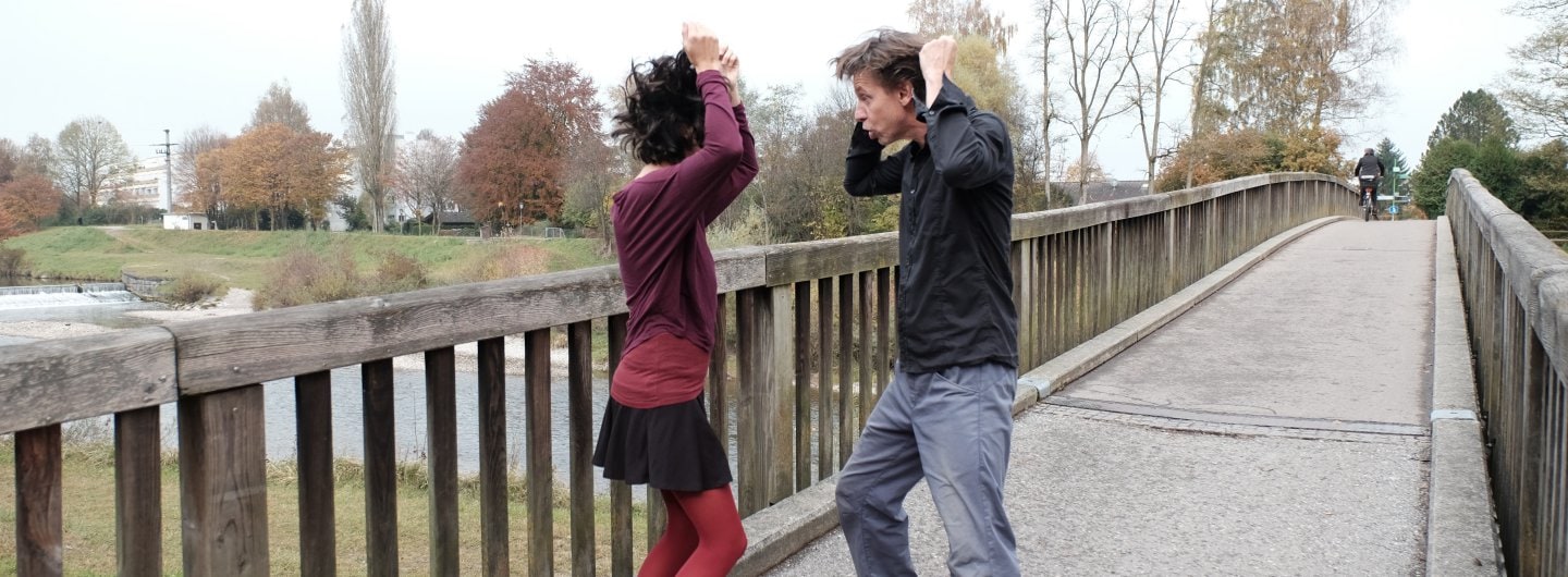 Paar steht voreinander auf einer Brücke und rauft sich tanzend die Haare