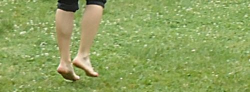 Eine grüne Wiese mit Gänseblümchen. Darauf ein paar nackte Füße, die gerade wieder auf der Wiese landen und in einer schwarzen Hose stecken die bis zum Knie geht